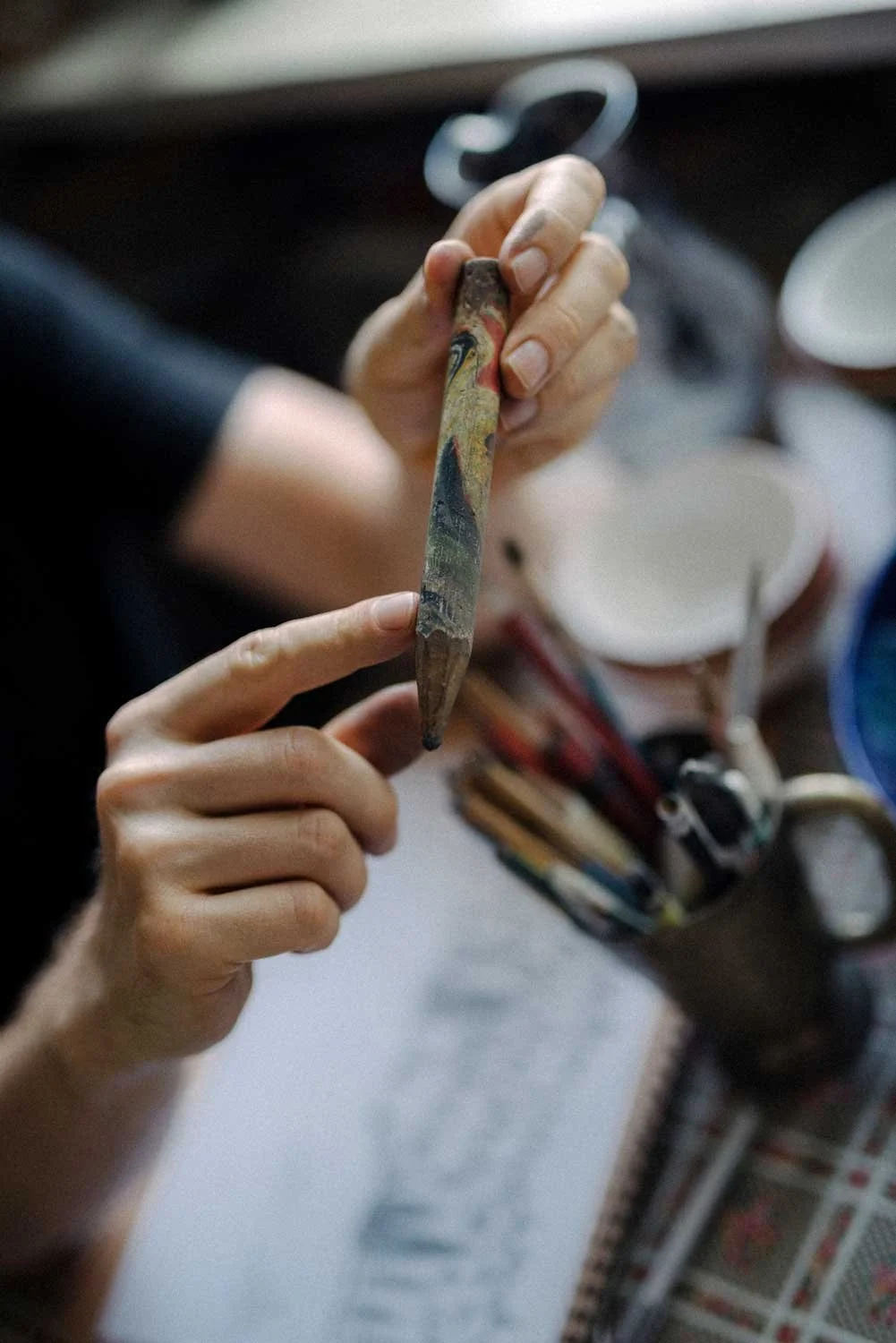 A person holding a paintbrush with a marbled handle, with other paintbrushes and a cup on a table in the background.