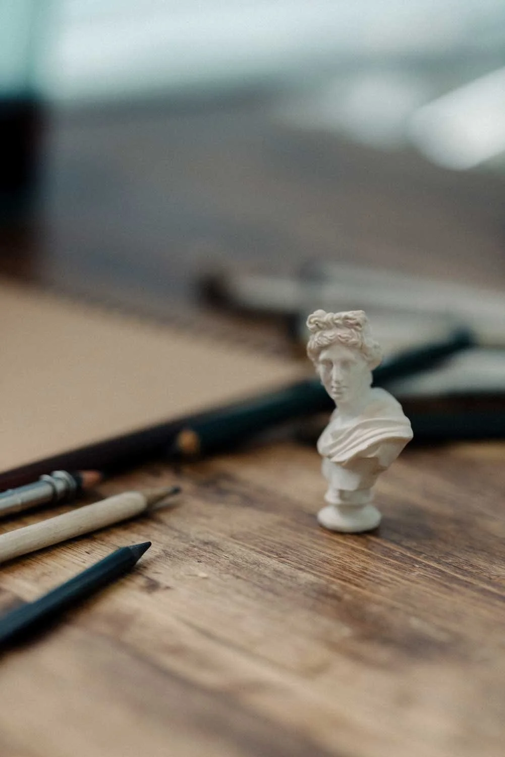 A small white bust sculpture of a woman's head and shoulders on a wooden desk. Art supplies, including pencils and pens, are scattered around the sculpture, with a notebook and blurred background.