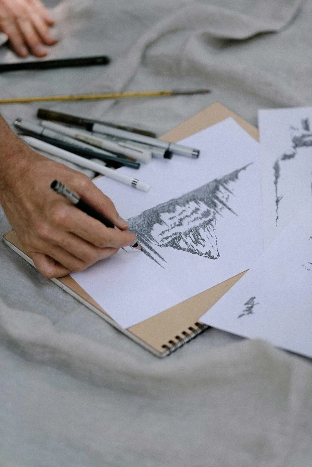 A person drawing a landscape scene with mountains and trees using a black marker on white paper, with several pens and pencils on the table.