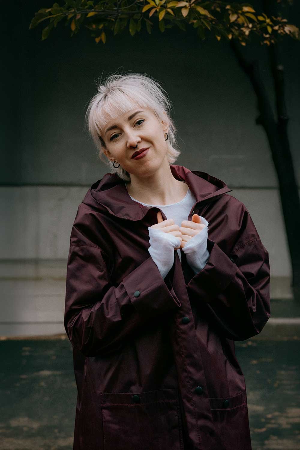 A young woman with short, platinum blonde hair and hoop earrings stands outdoors, wearing a dark red raincoat and white gloves, with her hands close to her chest, smiling gently at the camera.