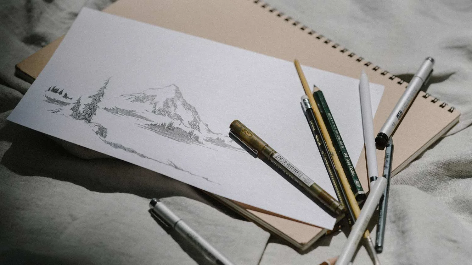 A sketchbook on top of two other notebooks with a detailed pencil drawing of a mountain and trees. Surrounding the sketchbook are various pens, pencils, and markers.