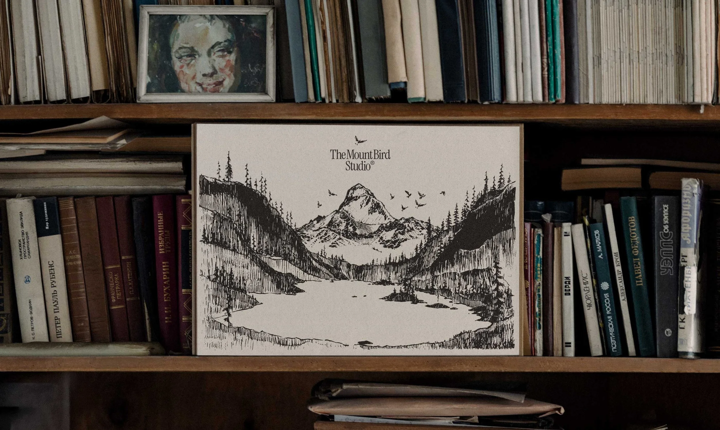 A bookshelf with books and a framed artwork. The artwork features a black and white sketch of mountains, trees, and birds, with the text 'The Mount Bird Studio'.