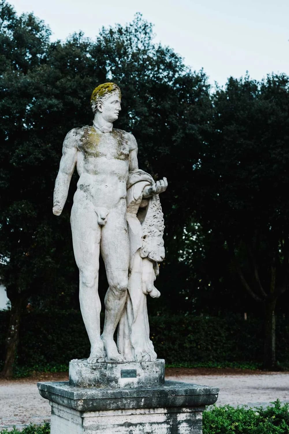 An outdoors marble statue of a male figure with a lush green tree background.