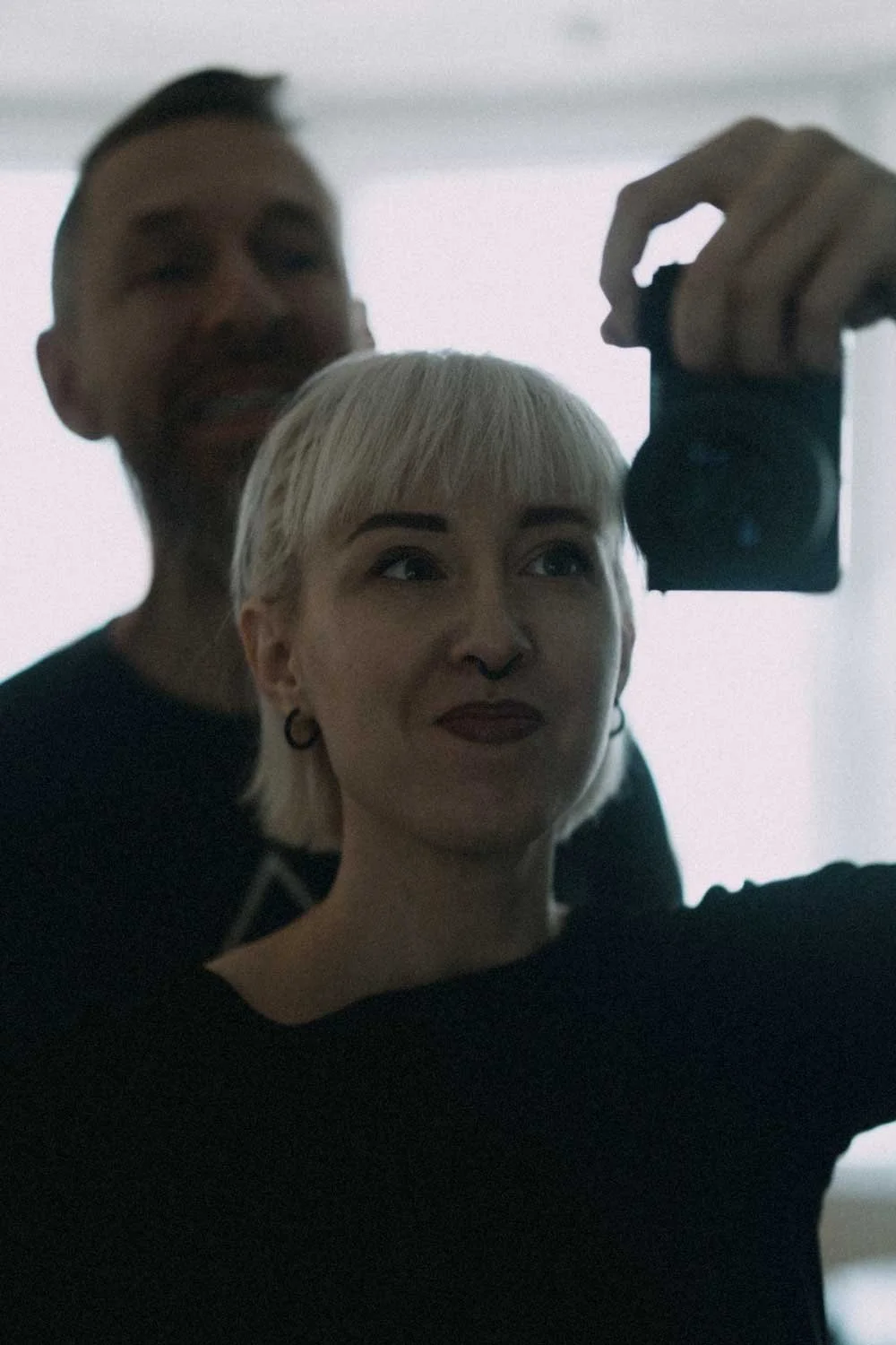 A person with short blonde hair and septum piercing taking a selfie in a mirror, with a man with dark hair and a beard standing behind them.