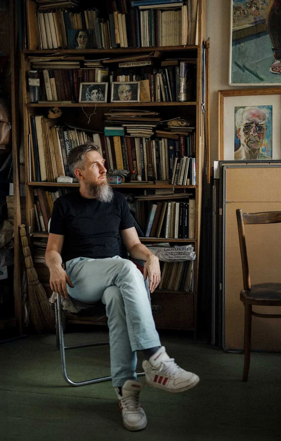 A man with a beard and black t-shirt is seated in a room filled with books and artwork, looking to his left.