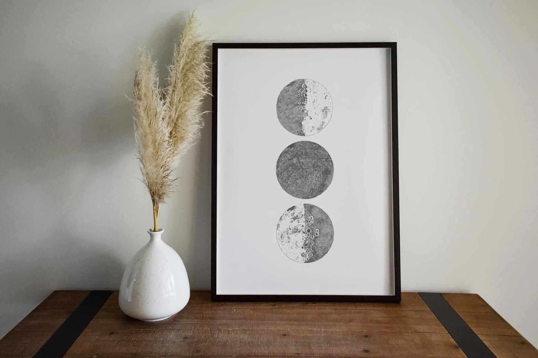 A white vase with pampas grass on a wooden table next to a framed black and white artwork of four moon phases on a plain wall.