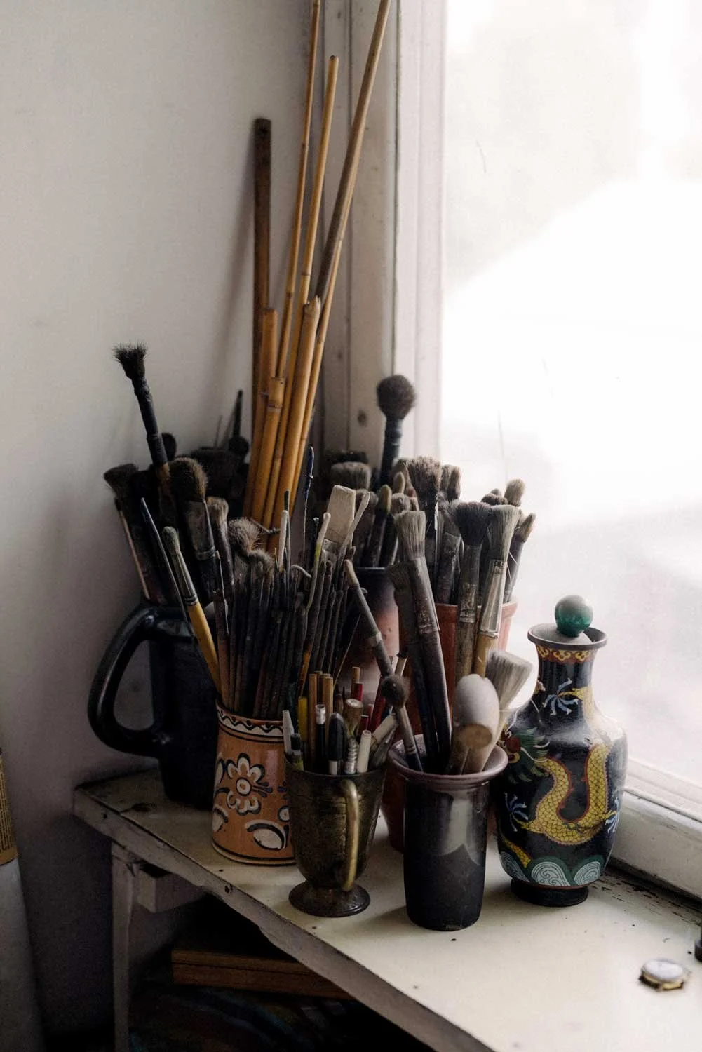 Collection of paintbrushes and artist tools in decorative vases and cups on a white table by a window.