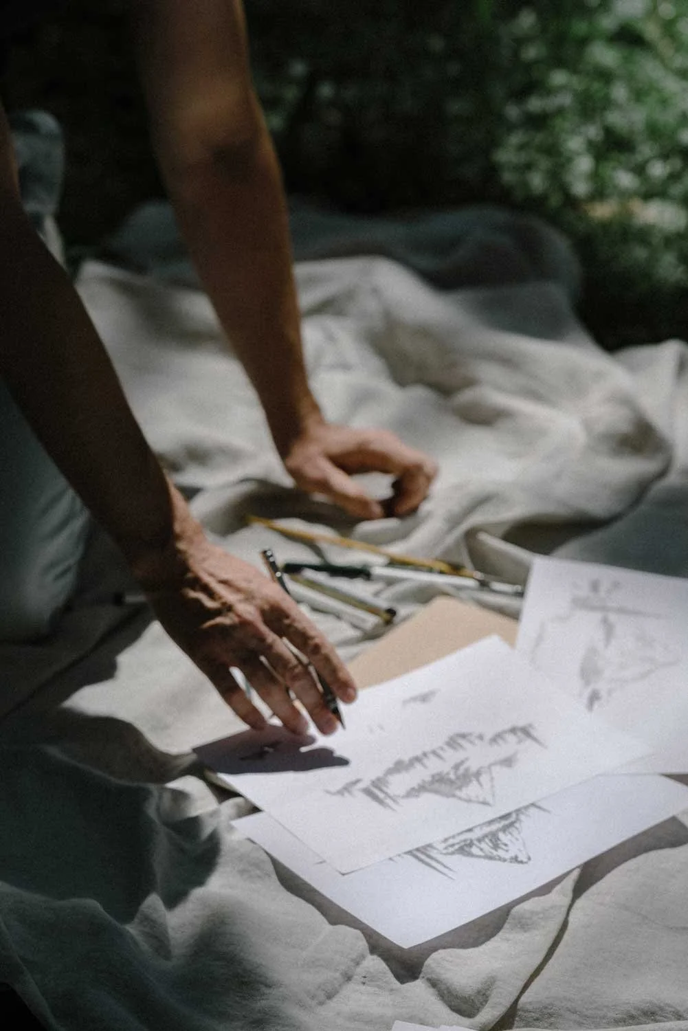 Person sketching on papers, surrounded by pencils, in a dimly lit environment with nature visible in the background.