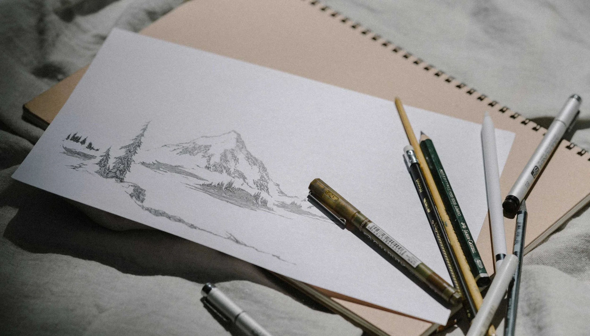 A sketchbook with a mountain landscape drawing, surrounded by pens and mechanical pencils, on top of two closed books or notebooks.