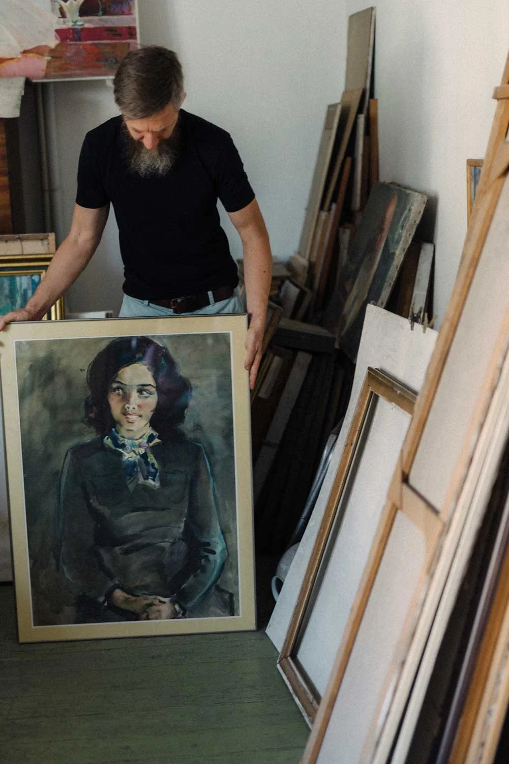 A man with a beard and gray hair looks down while holding a framed portrait of a woman with dark hair and a patterned scarf. The room is filled with various framed paintings and canvases leaning against the wall and stacked on the floor.