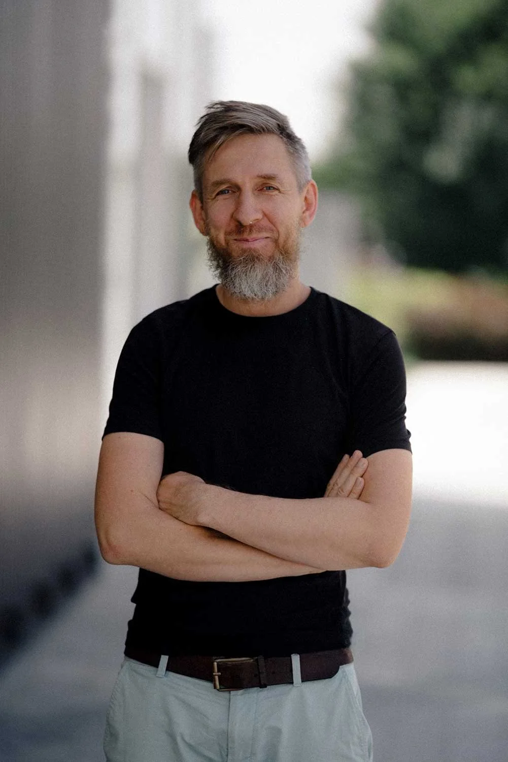 A middle-aged man with a beard, wearing a black t-shirt and light-colored pants, standing outdoors with arms crossed, smiling confidently.