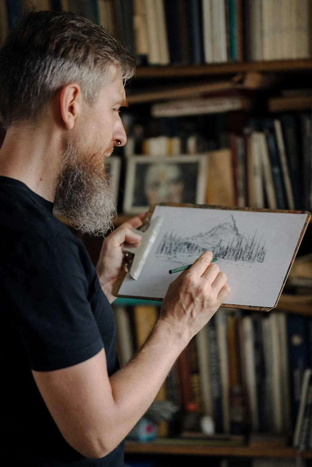 A man with a gray beard and short hair, wearing a black shirt, is drawing a mountainous landscape on a sketchpad in a room with bookshelves filled with books.