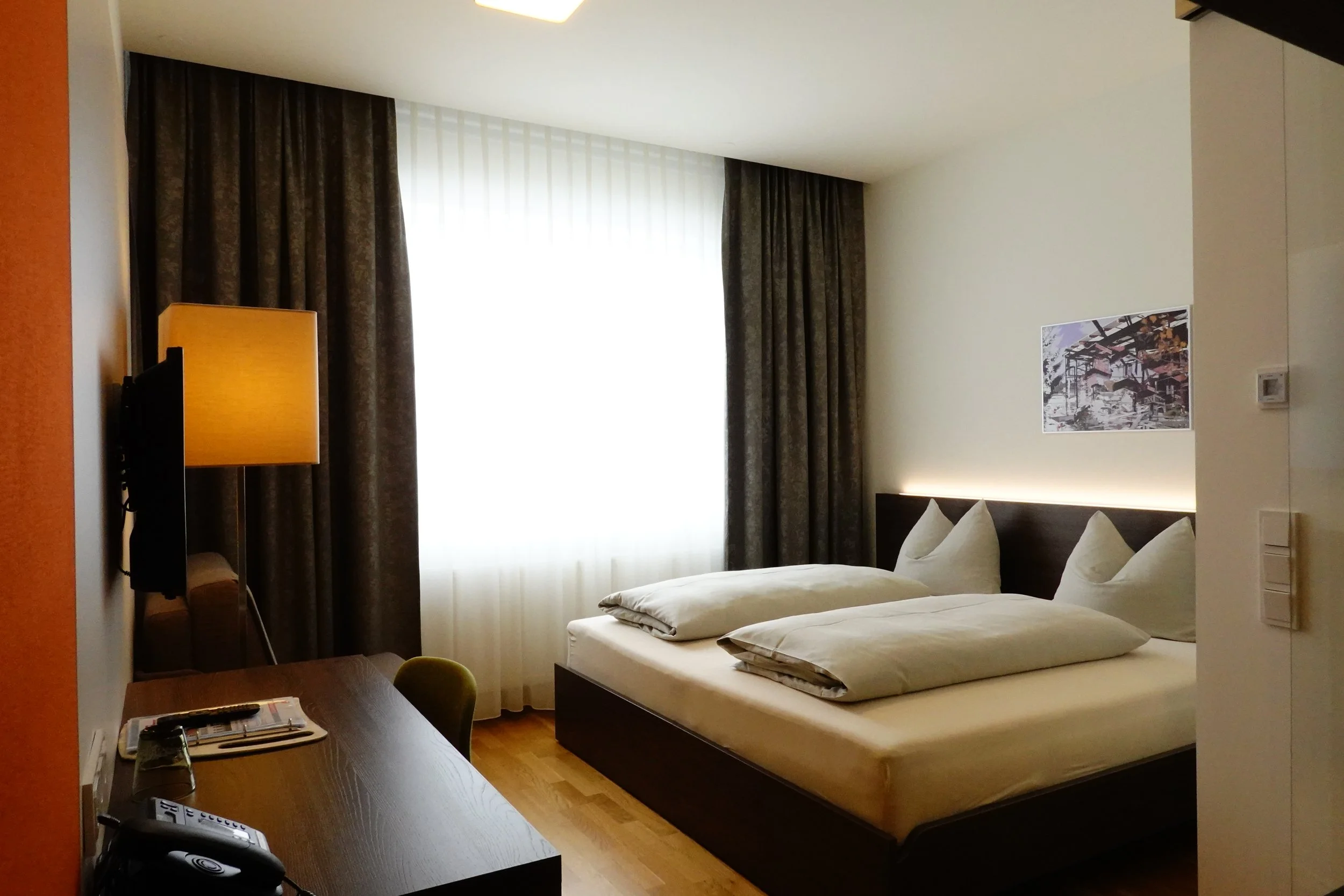 Ein modernes Hotelzimmer mit Arbeitsplatz, Fernseher, Doppelbett, kleinem Schreibtisch, Sessel. Der Raum ist hell und minimalistisch gestaltet.