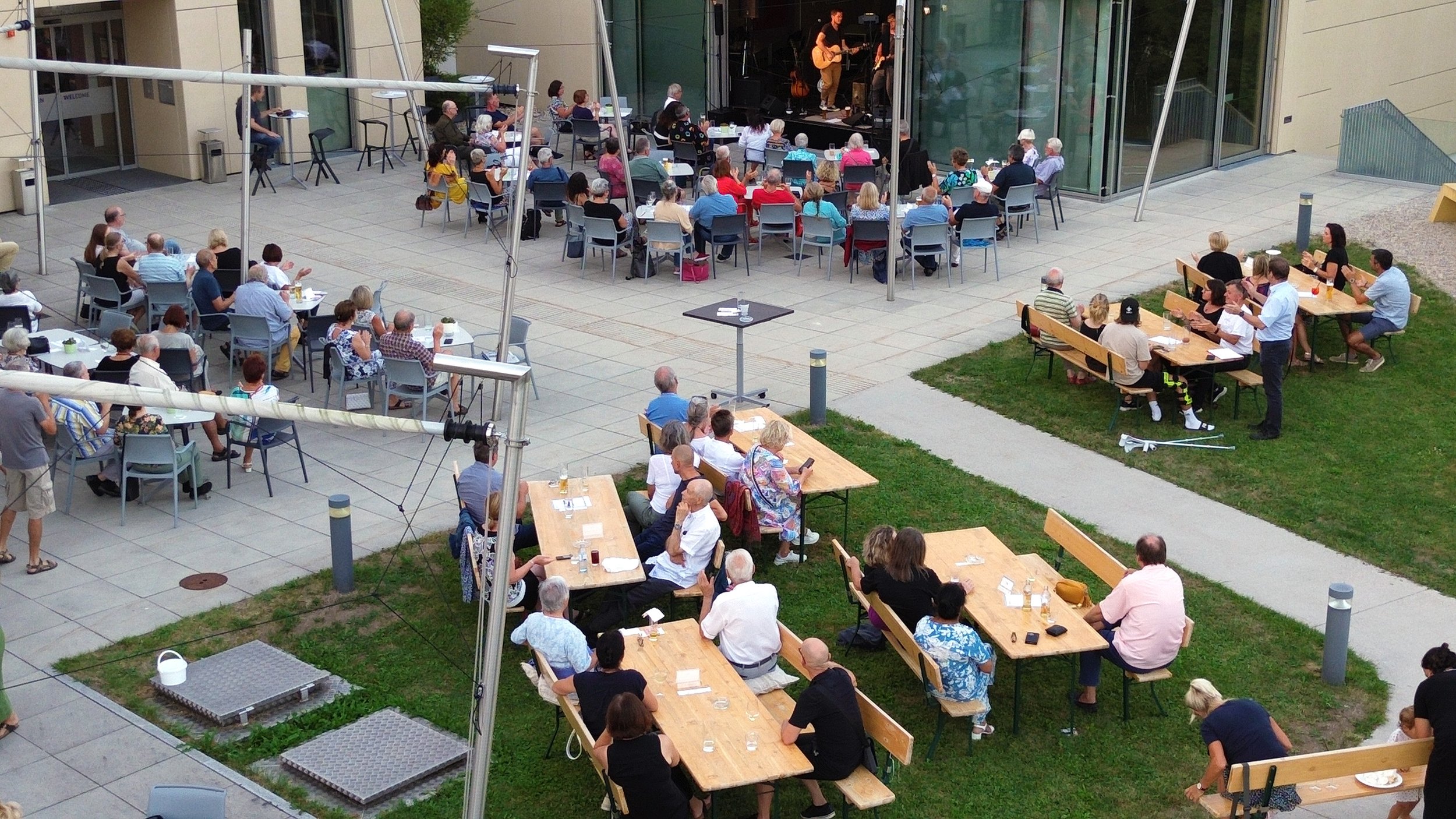 Menschen, die bei einem Konzert im Freien sitzen, mit Blick auf eine Bühne, auf der Musiker auftreten. Einige sitzen an Tischen, andere auf Bänken, alles auf einer gepflasterten Fläche zwischen Gebäuden.