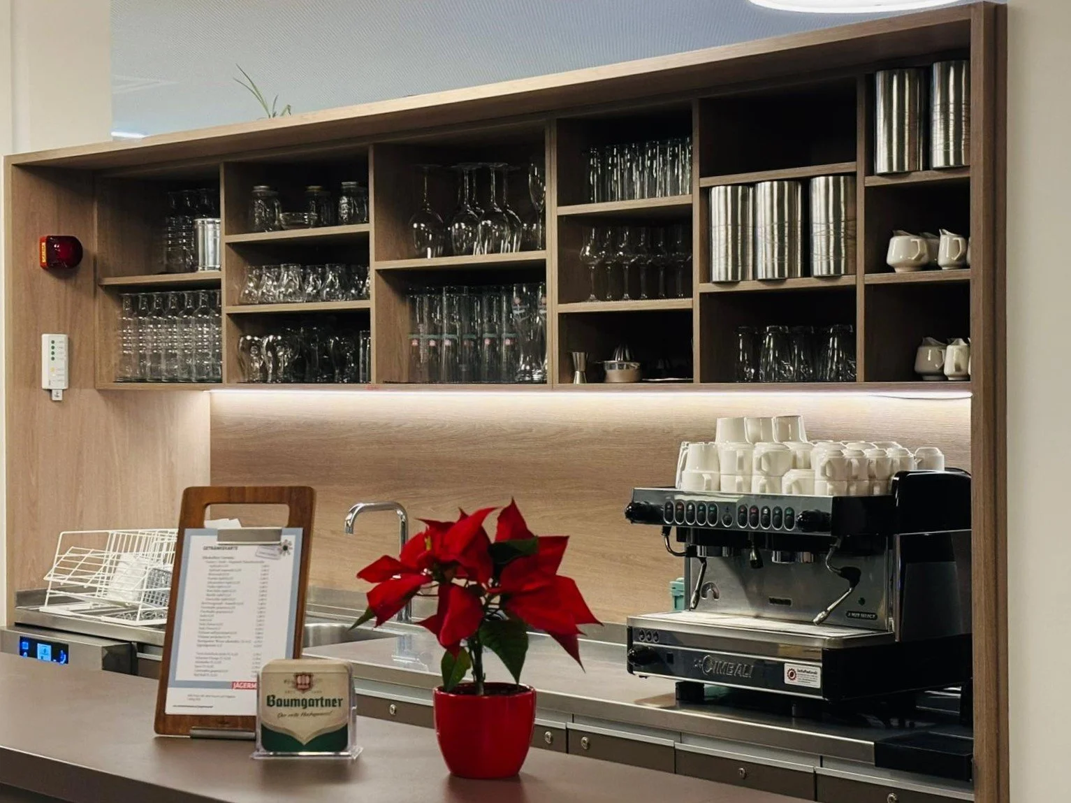 Kleine Bar- oder Theke-Ecke eines Cafés mit einer Espressomaschine, einem roten Weihnachtsstern in Topf, offenem Menü, Gläsern im Regal und einer Geschirrablage.