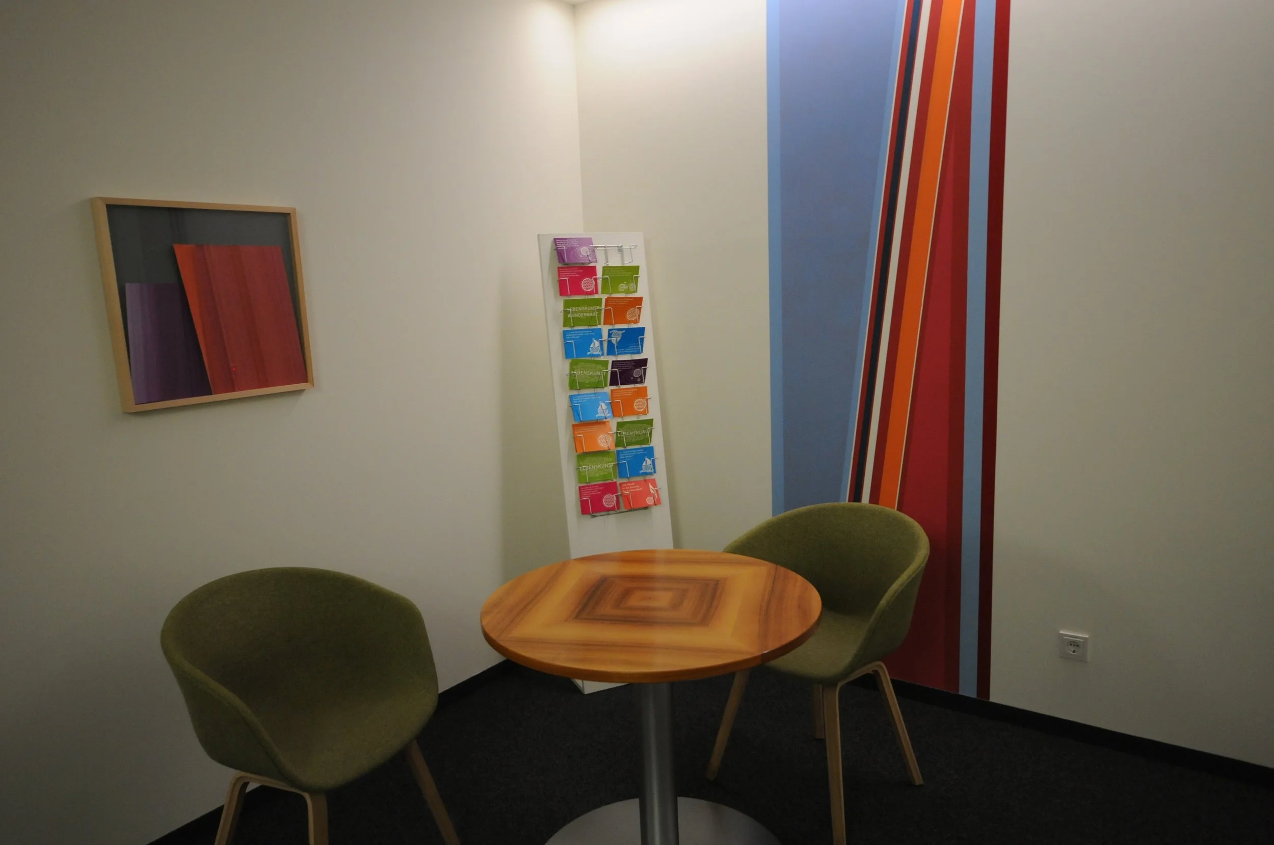 Ein kleines Wartezimmer mit einem runden Holztisch, zwei grünen Stühlen, einer Wand mit farbigen vertikalen Streifen und einer Wand mit einem gefassten abstrakten Kunstwerk.