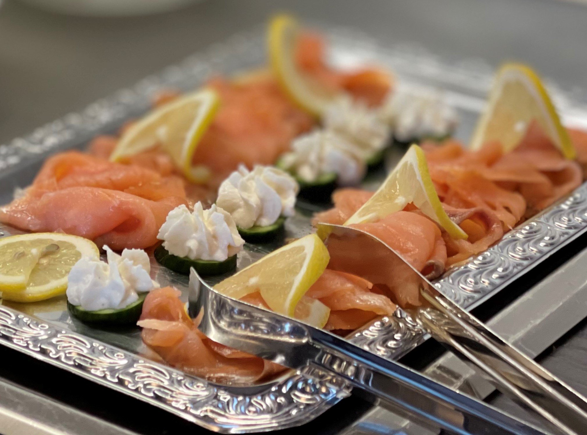 Kalte Fischplatte mit gehobeltem Lachs, Zitronenscheiben, Limetten mit Schlagsahne, Gurkenscheiben und Toppings auf einem silbernen Servierteller.
