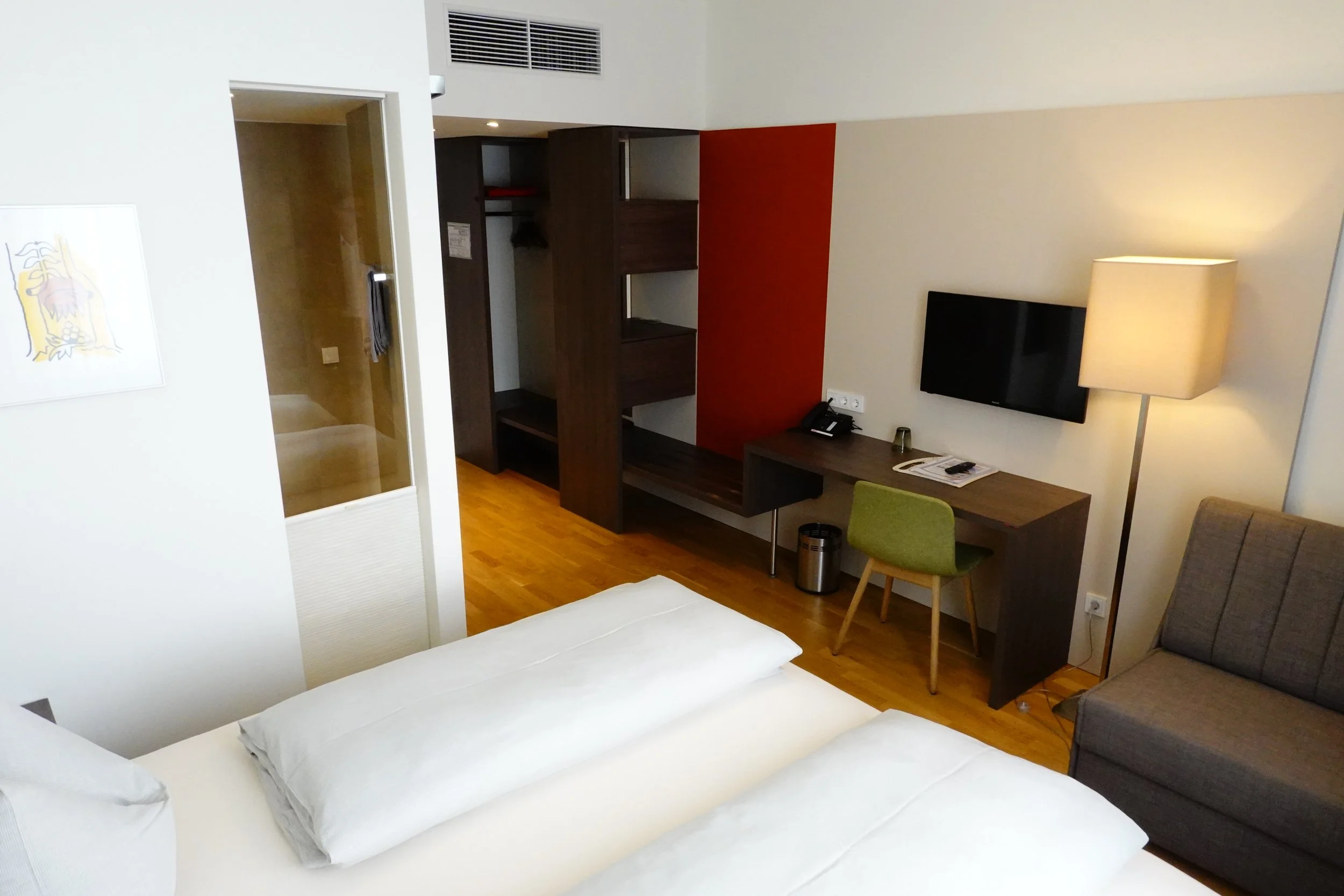 Ein modernes Hotelzimmer mit Arbeitsplatz, Fernseher, Doppelbett, kleinem Schreibtisch, Sessel, Schrank und Bad. Der Raum ist hell und minimalistisch gestaltet.