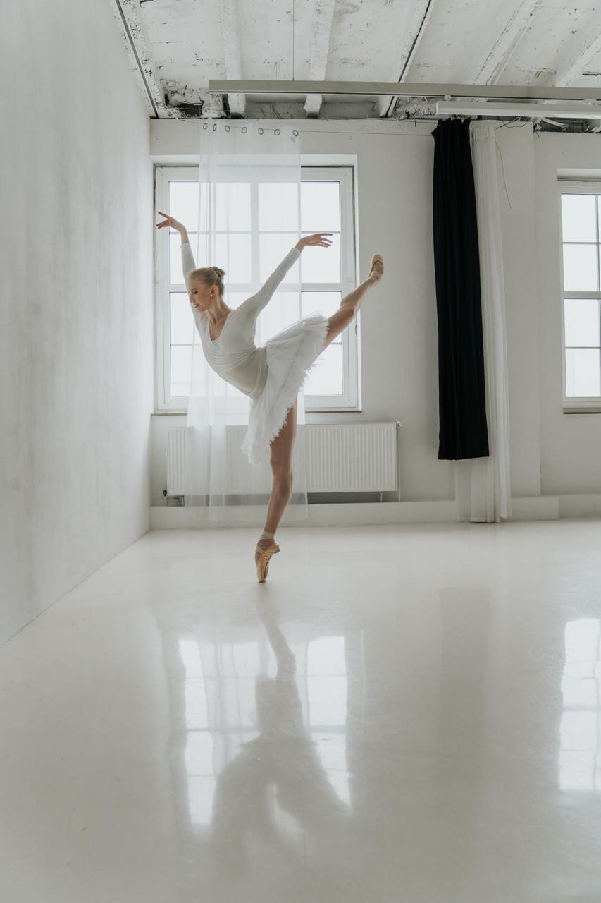 A ballerina dressed in white ballet attire is performing an arabesque pose on pointe in a bright, minimalistic dance studio with large windows and black and white curtains.