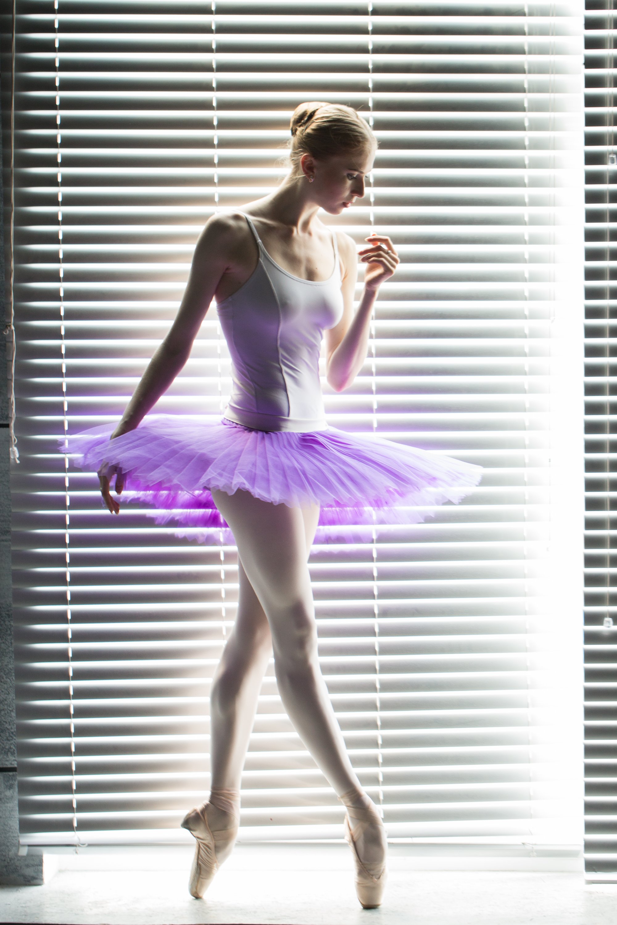 A ballerina in a purple tutu and ballet slippers stands in front of window blinds, striking a pose.