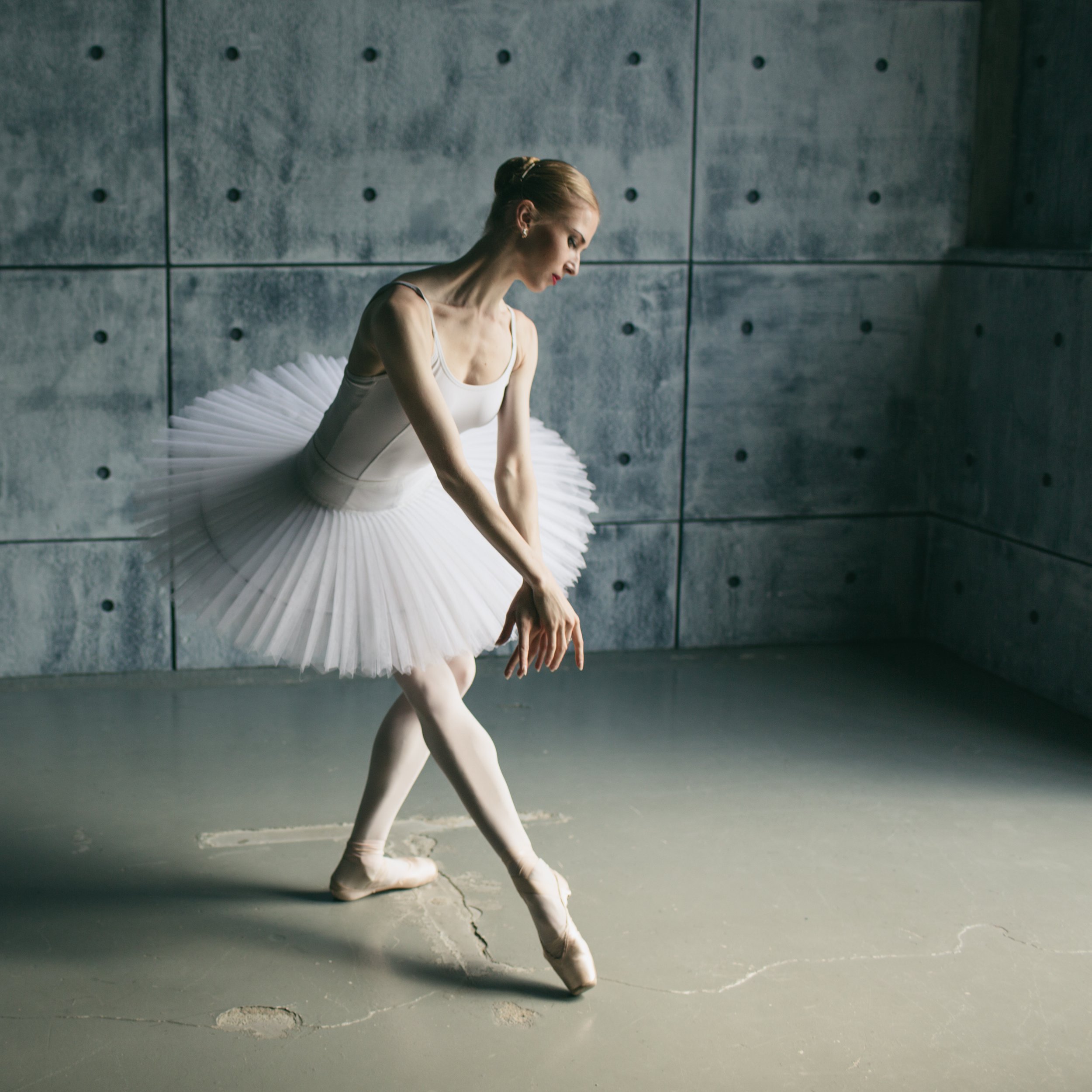 A ballet dancer in a white tutu standing on tiptoe on a cracked concrete floor.