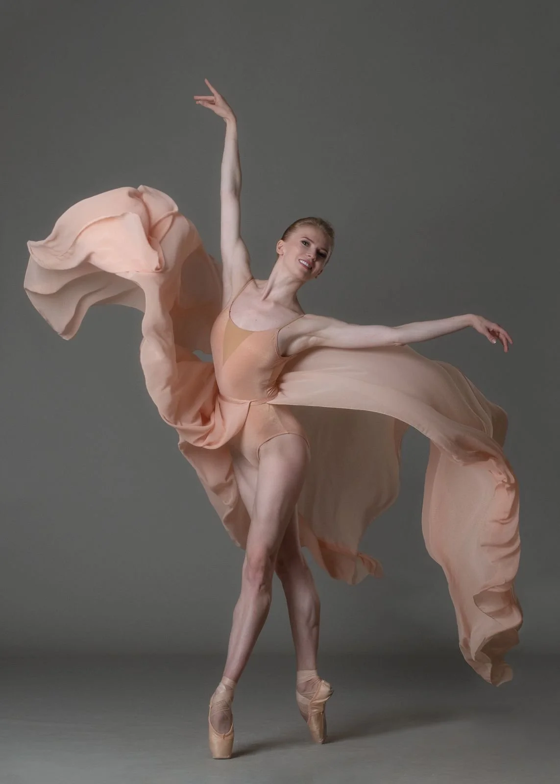 Ballet dancer mid-performance in a peach-colored flowing dress on pointe against a gray background.