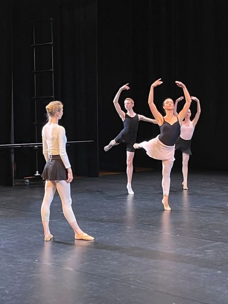 Four ballet dancers rehearsing on stage, with three of them in ballet poses and one standing and watching.