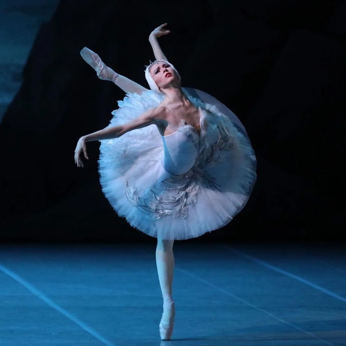 Ballet dancer performing en pointe in a white tutu with intricate embroidery, with one arm raised above her head and the other extended to the side, against a dark stage background.