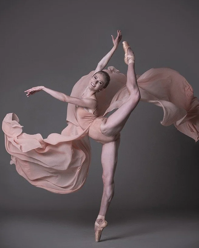 Ballet dancer in a flowing peach-colored dress performing a high leap on pointe with a gray background.