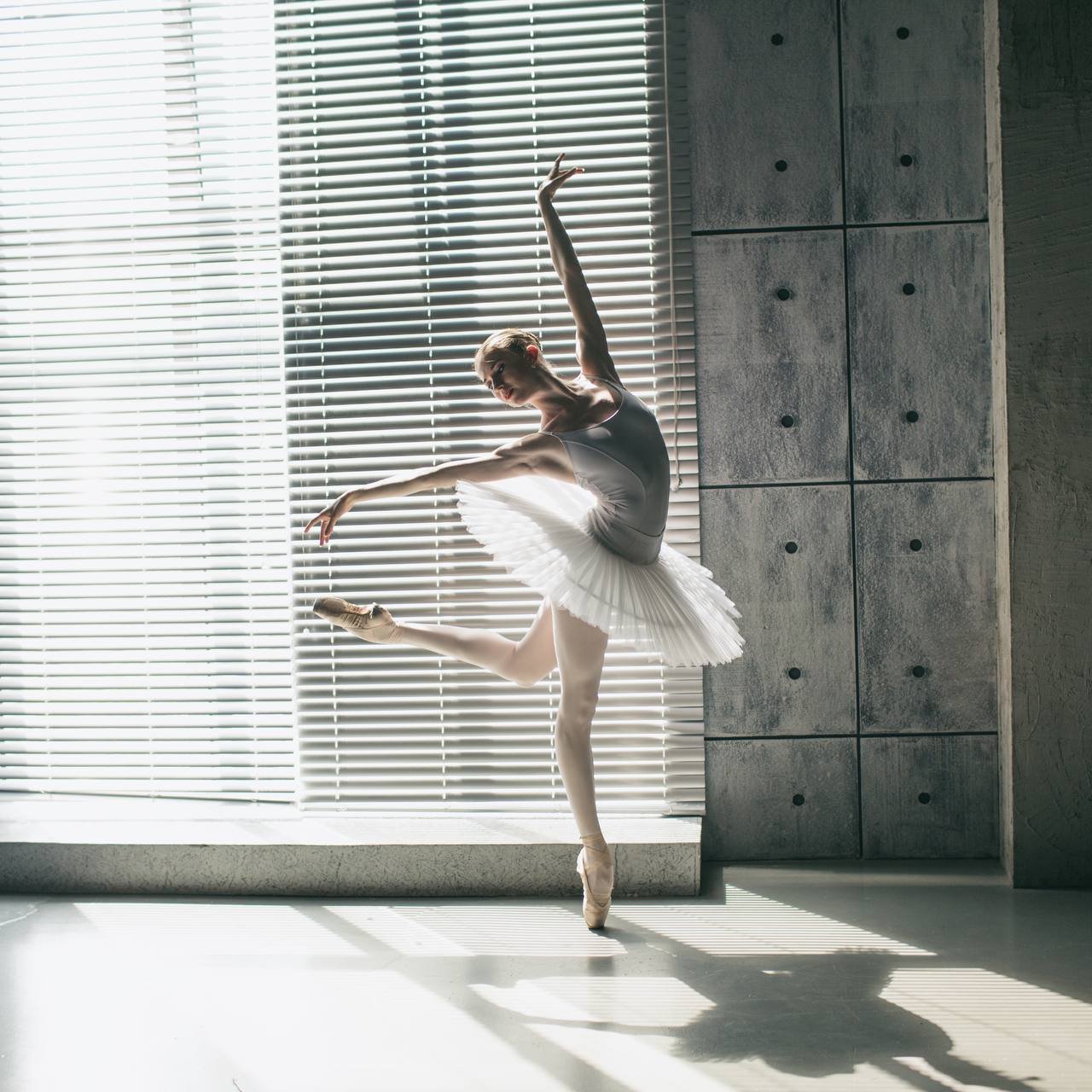 Ballet dancer practicing near a window with blinds in a modern studio.