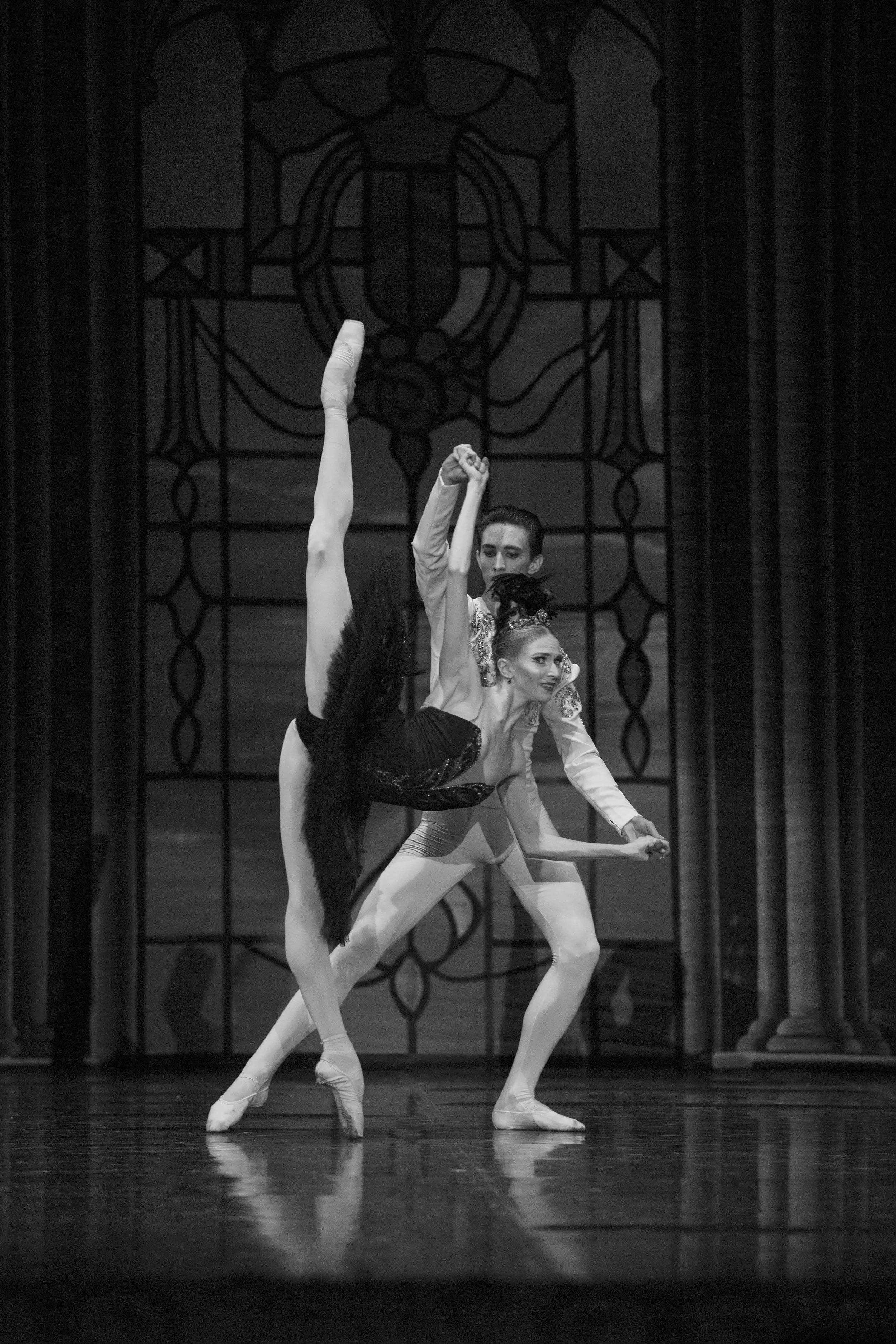 Ballet dancers performing on stage in front of a stained glass backdrop, with one dancer performing an extended leg move while the other supports.