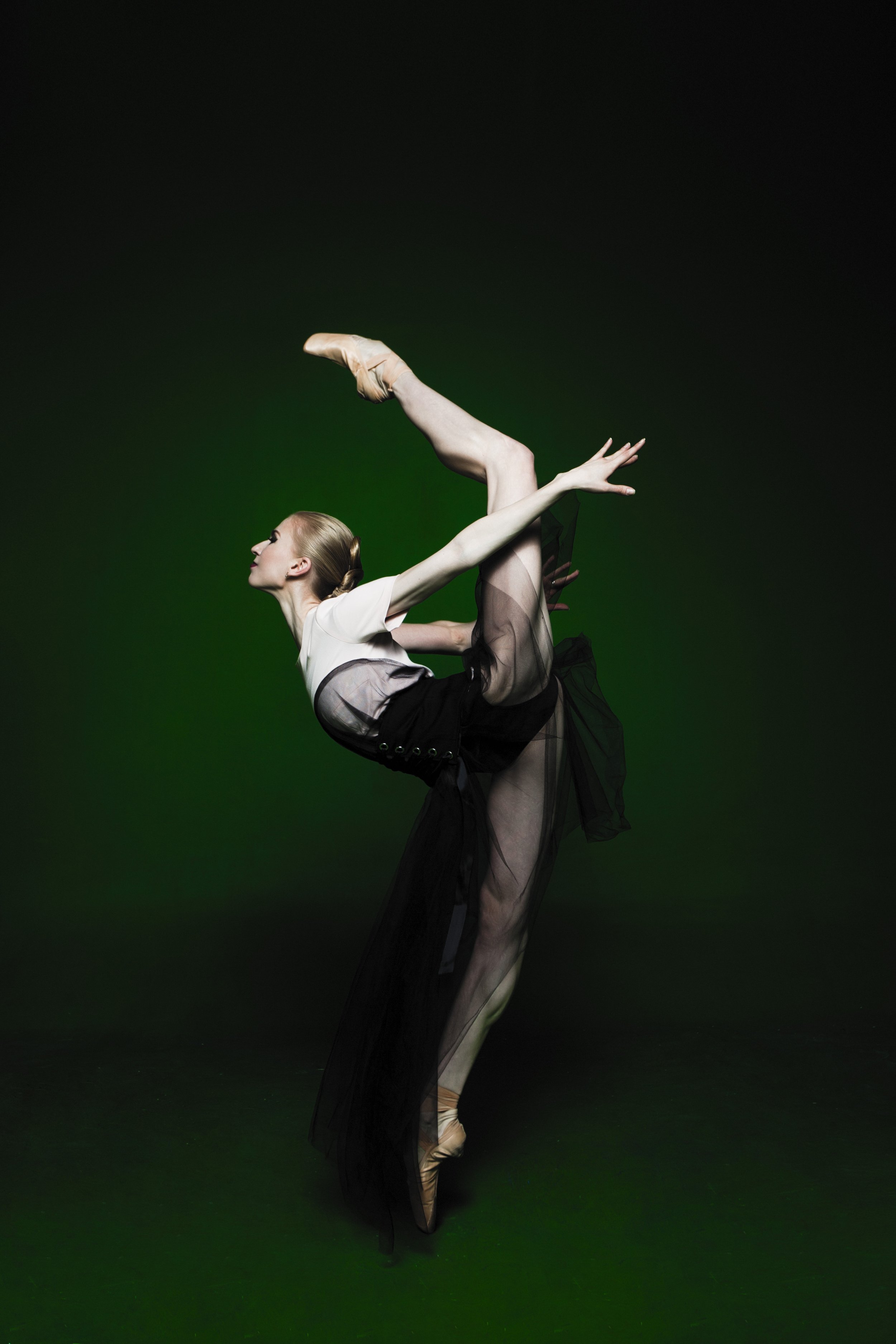 Ballet dancer performing a pose in front of a dark background with green towards the bottom.