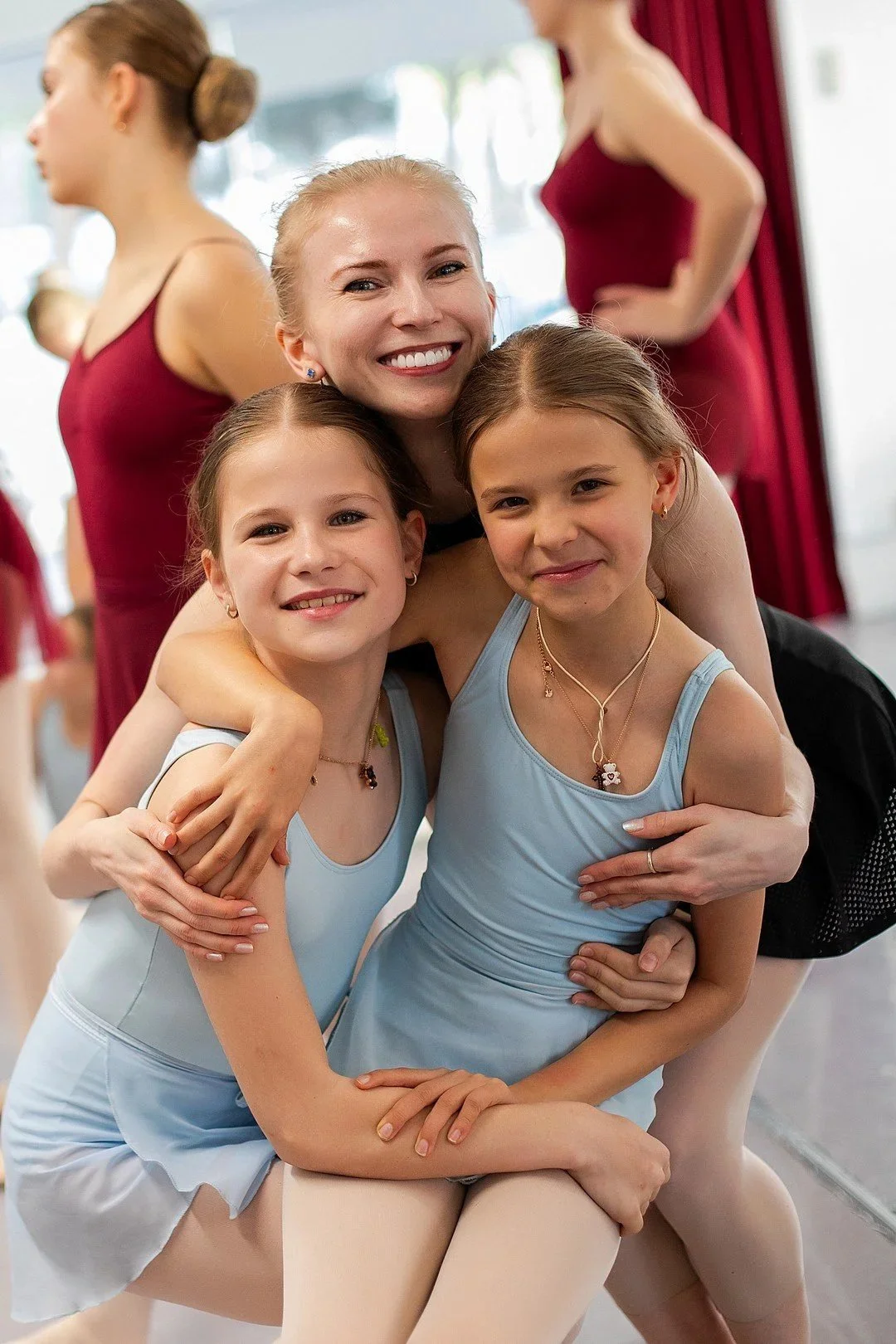 Two young girls in blue leotards and tights smiling as they are embraced by a smiling adult woman, likely their coach, in a dance studio with other dancers in the background.