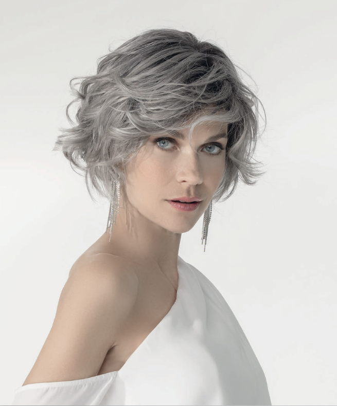 Portrait of a woman with short, wavy, gray hair, blue eyes, wearing long dangling earrings and a white off-shoulder top against a plain background.