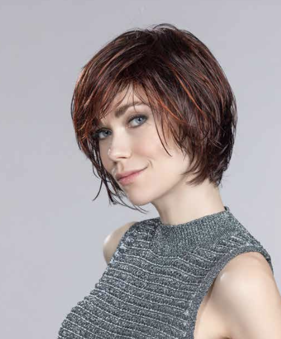 A woman with short, layered, reddish-brown hair and blue eyes, wearing a sparkly gray sleeveless top, smiling subtly against a plain gray background.