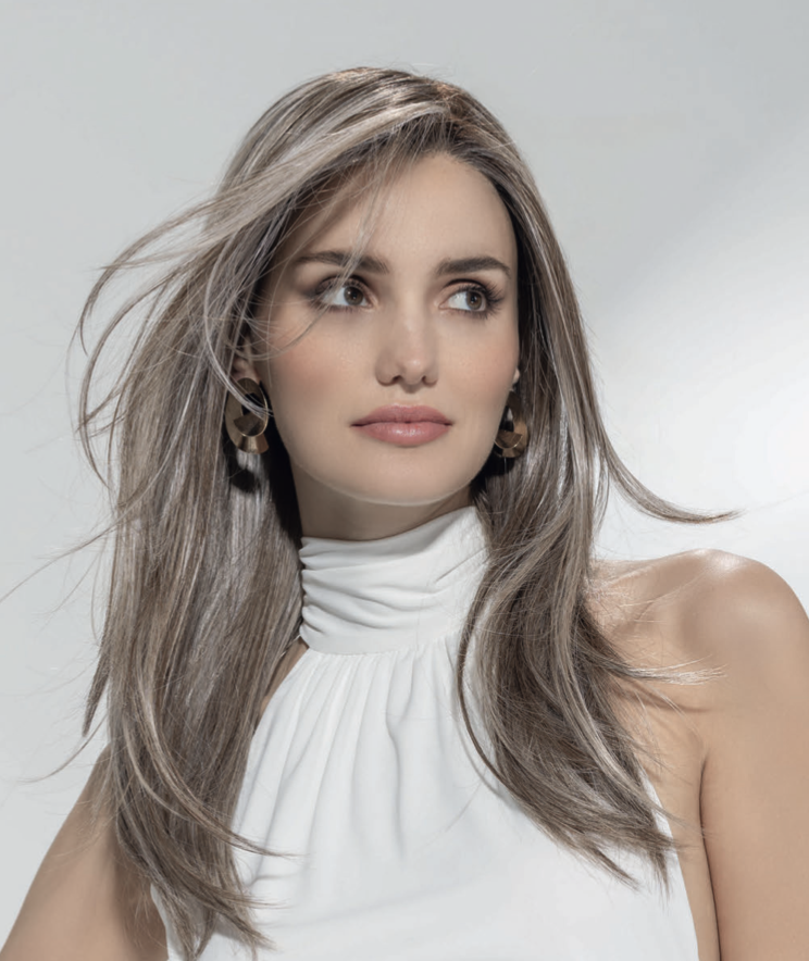 A woman with long, light brown hair and light skin looking to the side, wearing a white high-neck top and earrings, against a plain light background.