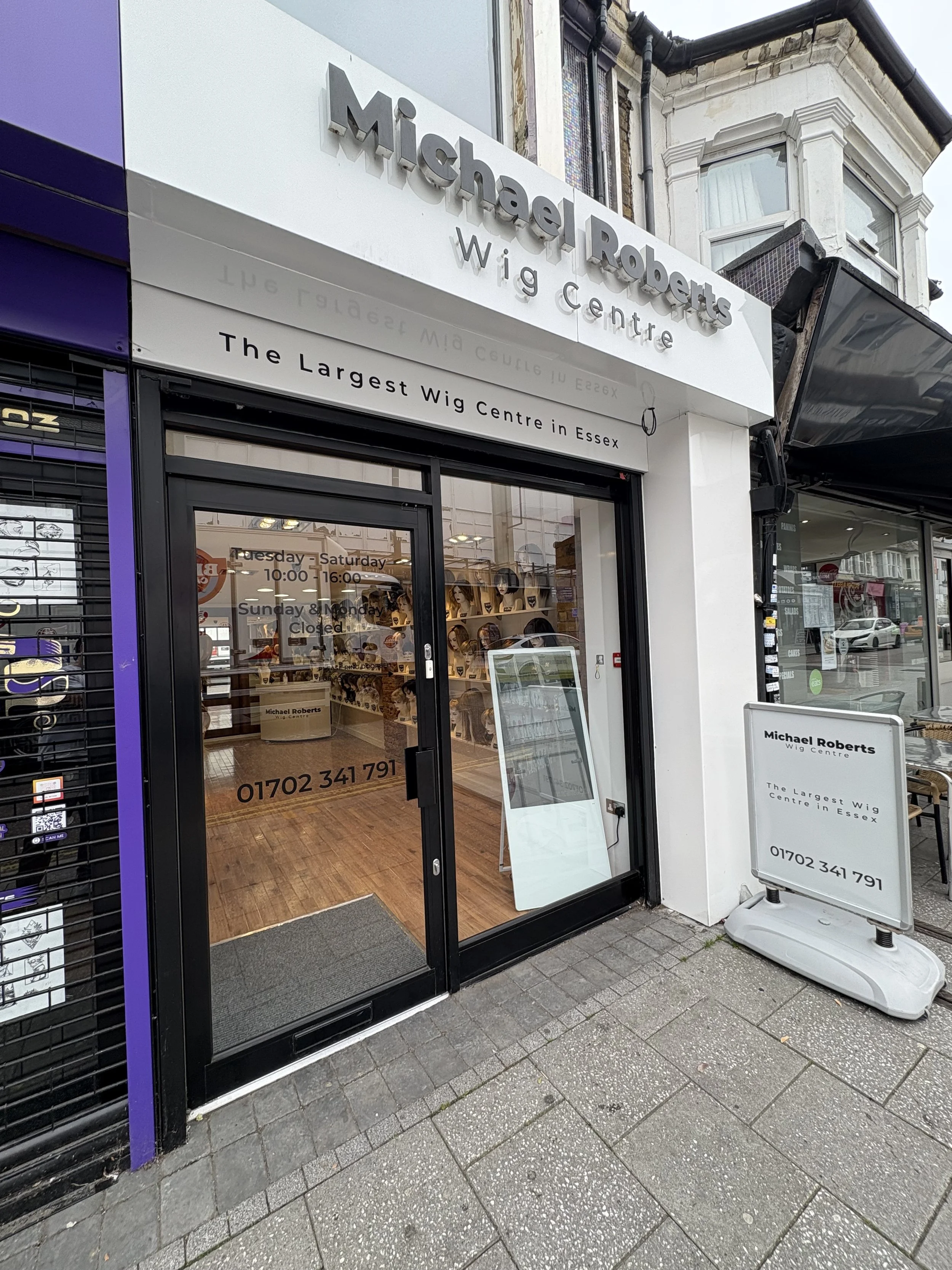 The storefront of Michael Roberts Wig Centre, a wig shop in Essex, featuring a white sign with black text, large glass doors with operating hours, a stand with a menu of services outside, and through the glass, displays of wig mannequins and hairpieces inside.