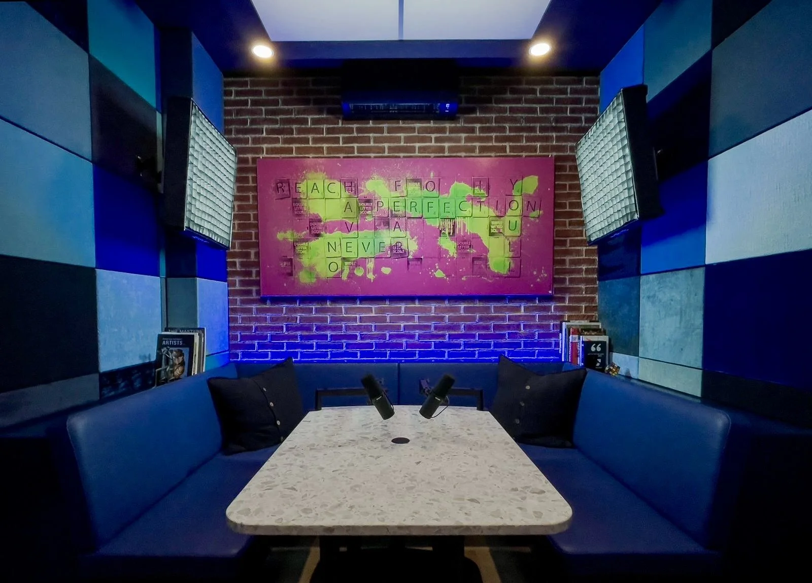 A television screen shows video editing or live streaming software in a studio with a pink background and green text on the wall behind, with an additional computer monitor on the right and various control devices in the foreground.