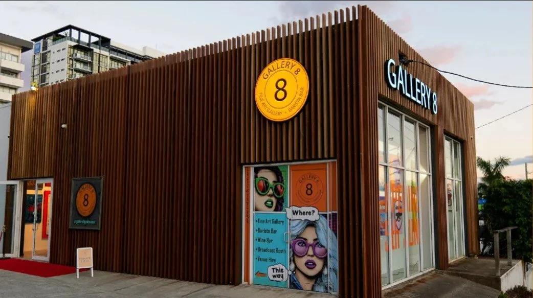 Exterior view of Gallery 8 building with wooden slat facade, signage on the wall, and colorful poster featuring two illustrated women with sunglasses in front of a sunset background.
