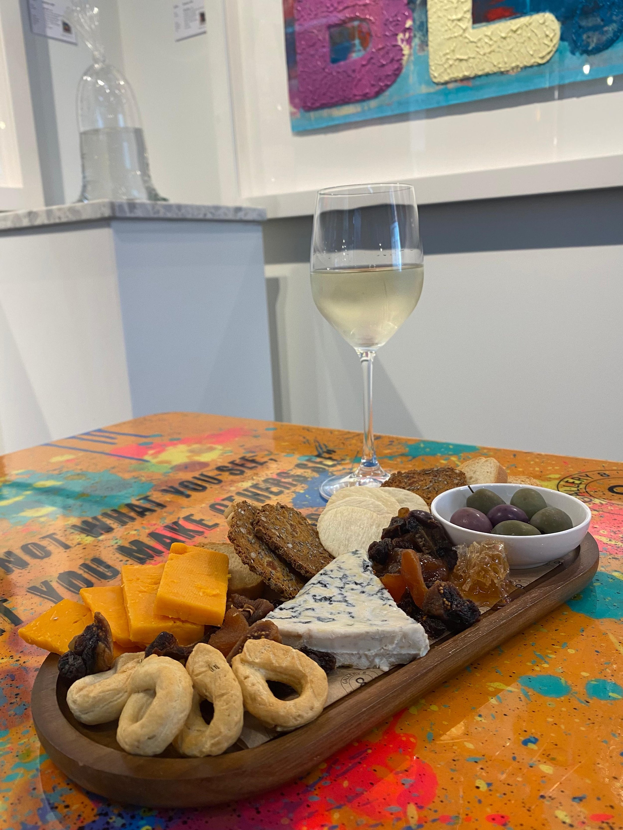 A wooden serving board with assorted cheeses, crackers, dried fruits, olives, and a glass of white wine on a colorful table with a glass water bottle in the background.