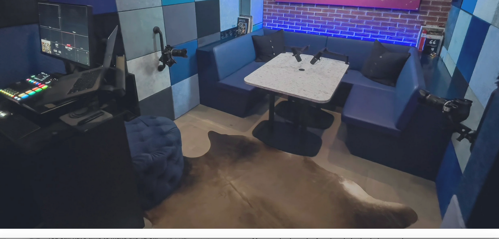 A video production setup with a monitor showing a control interface, a laptop, and a wall-mounted TV displaying a colorful art piece with the words "REACH FOR A PERFECT NEVER QUIT" against a pink background with green paint splatters. The room has brick walls and soundproofing panels in blue, gray, and black.