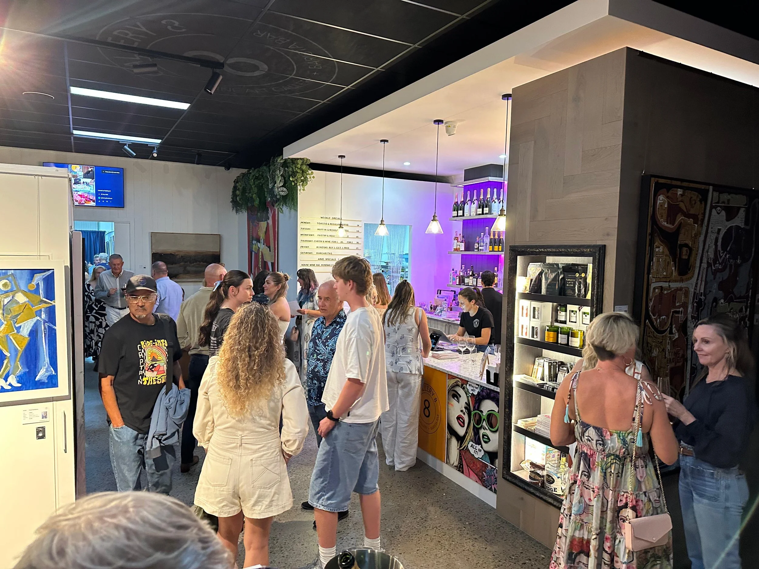 People gathered inside a bar or cafe, chatting and waiting in line. The decor includes a bar shelf with bottles, artwork on the walls, and hanging pendant lights.