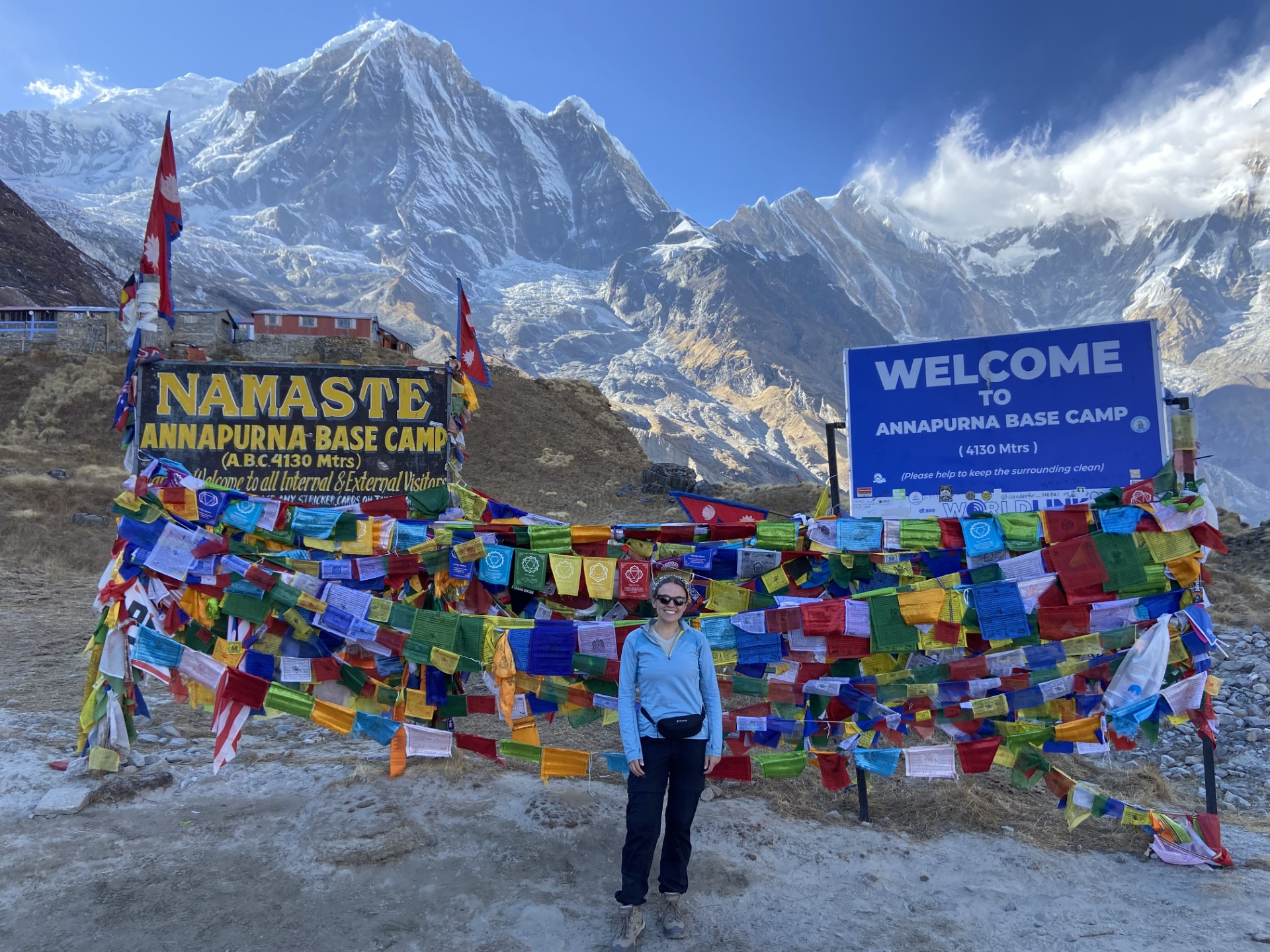 Georgia is standing smiling and wearing sunglasses in front of lots of muticoloured bunting and two large signs that say 'Welcome to Annapurna Base Camp' and 'Namaste'. Behind her, a range of the Himalayan mountains is illuminated in sunlight