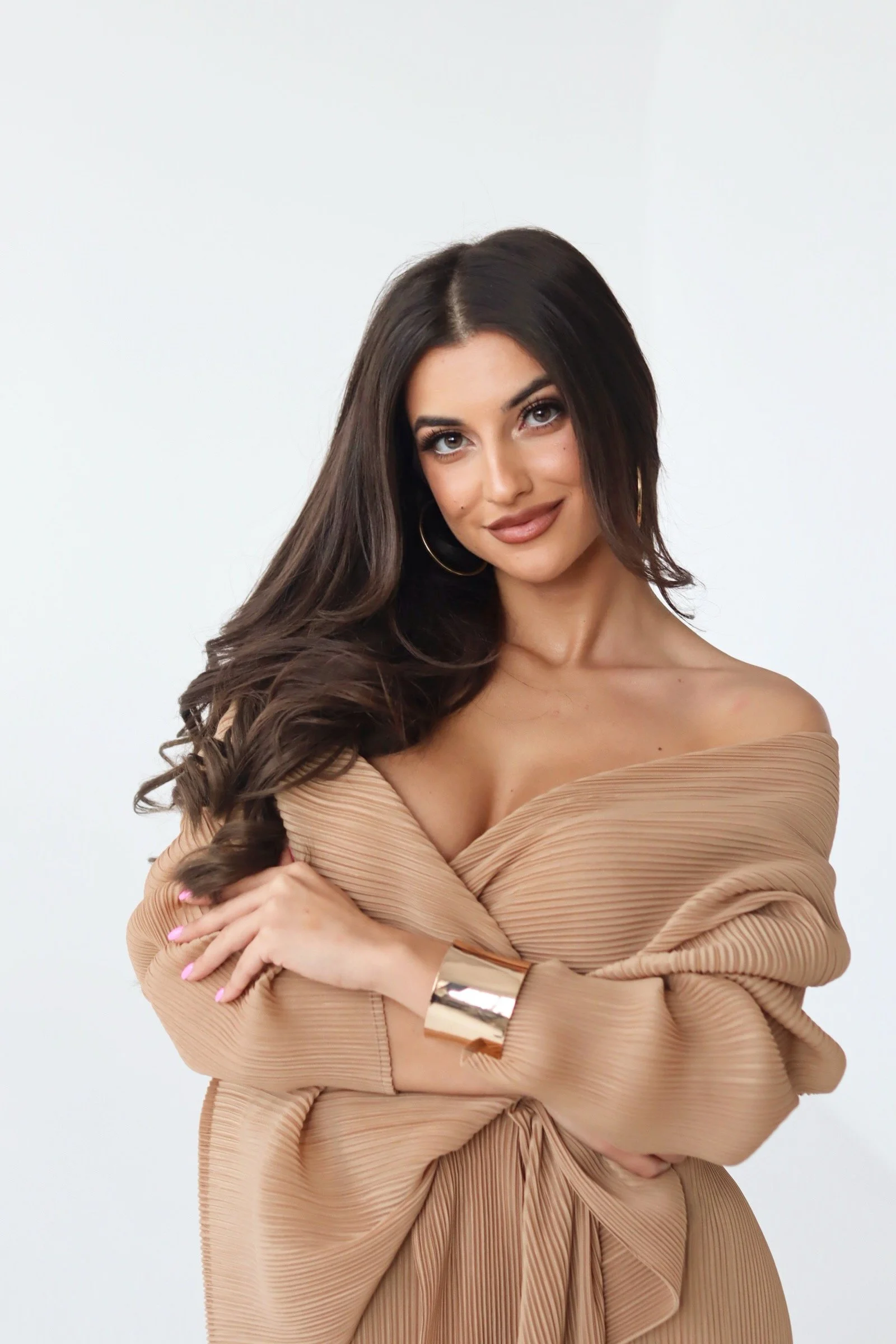 A woman with long dark hair, gold hoop earrings, and a warm smile, posing against a white background, wearing an off-the-shoulder beige pleated dress, with her arms crossed.