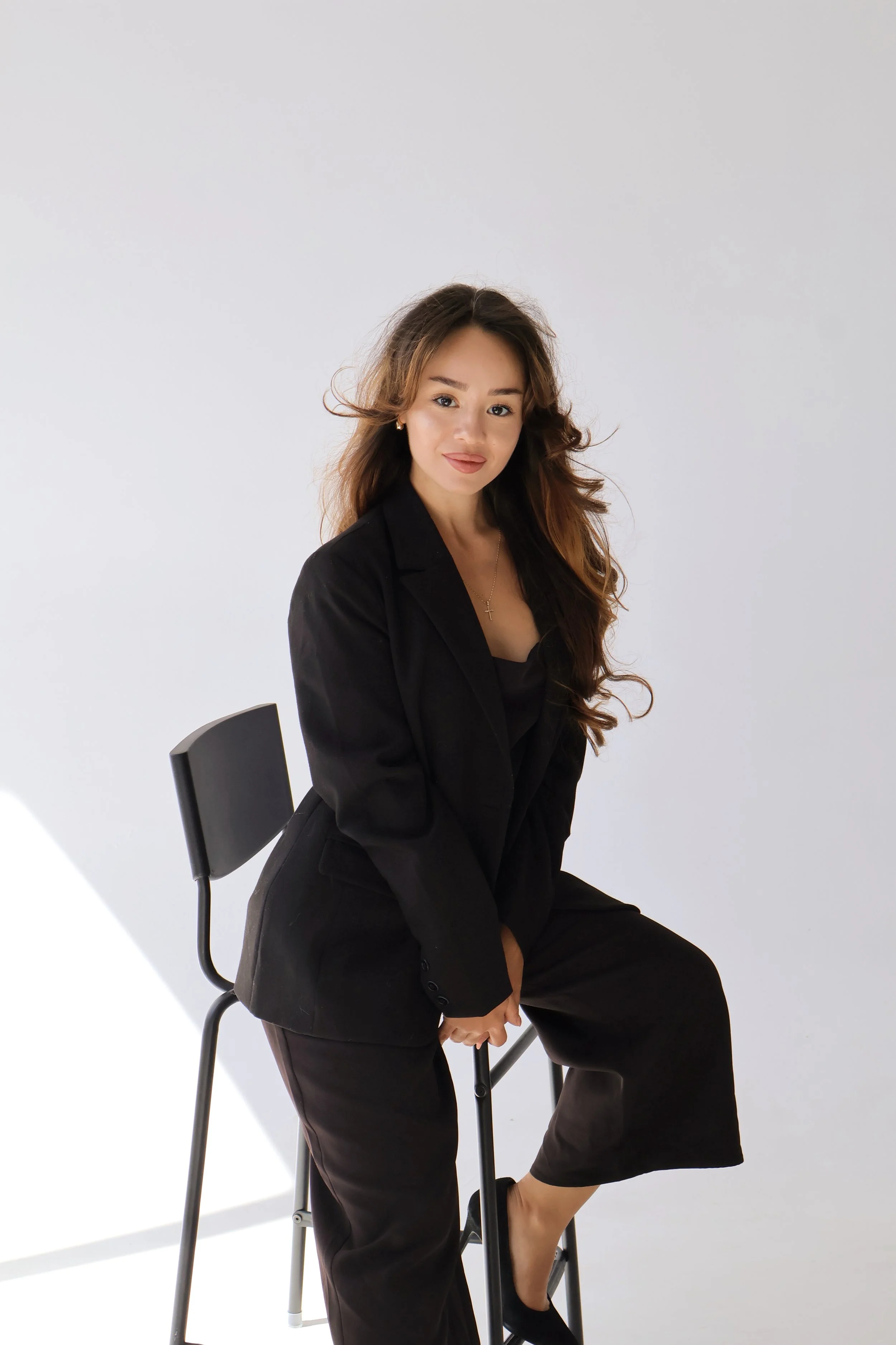A woman with long, wavy brown hair sitting on a black chair with a plain white background. She is dressed in black and looking at the camera.