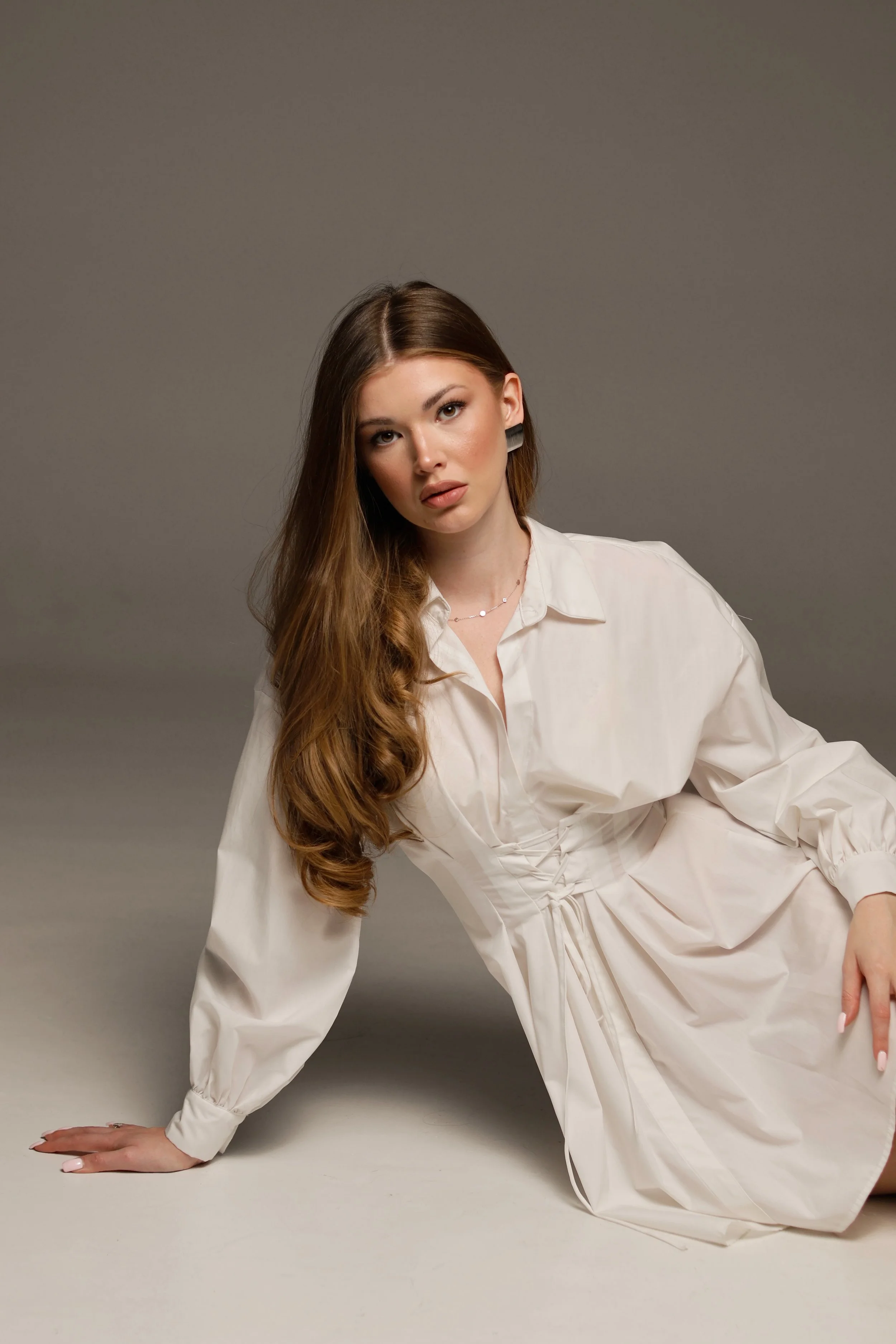 A woman with long, wavy brown hair, wearing a white dress, is posed on the floor against a plain gray background. She is supporting herself with one hand on the ground and has a neutral expression.