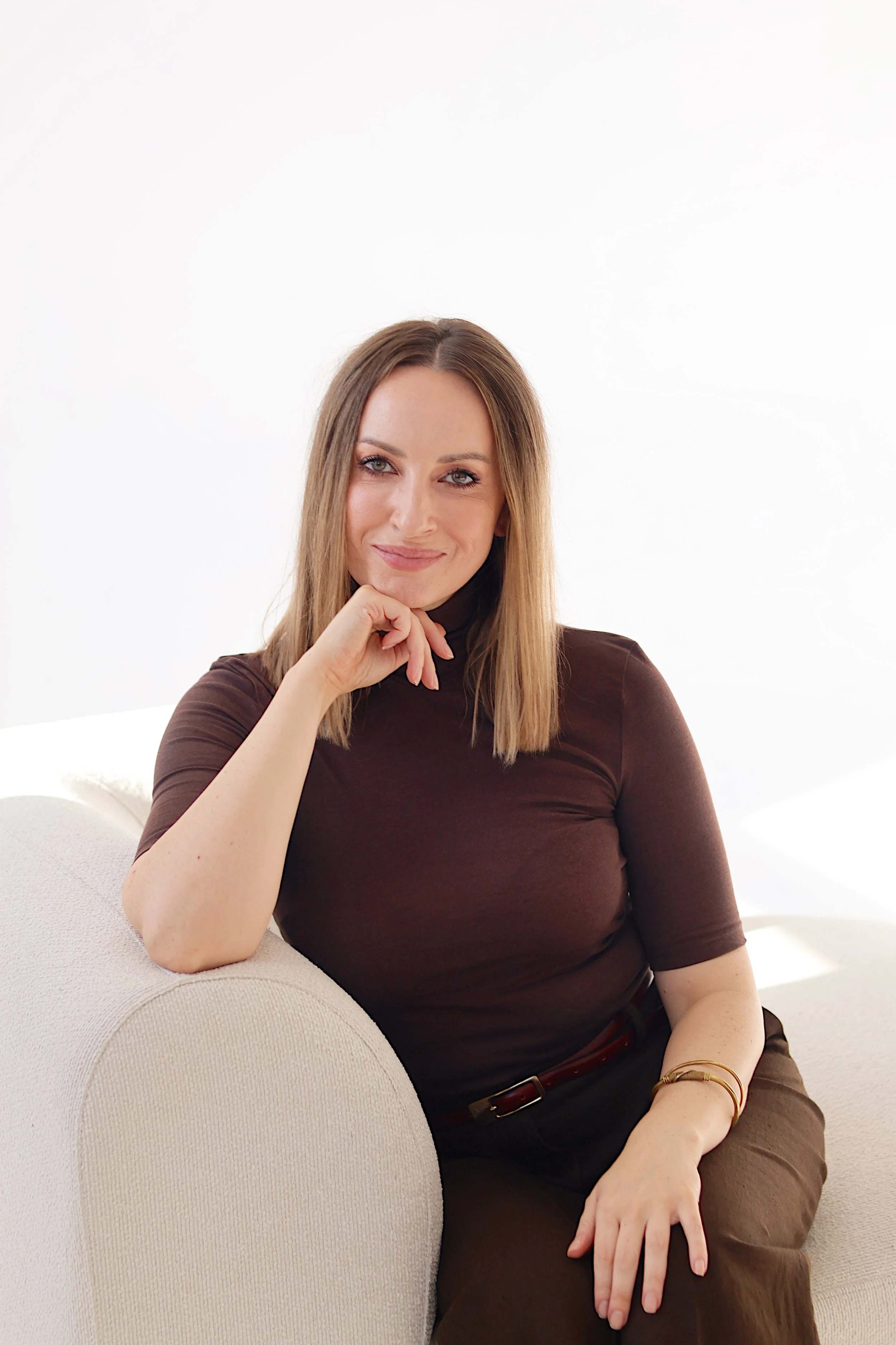 A woman with straight, shoulder-length blonde hair, wearing a brown shirt and brown pants, sitting on a light-colored sofa with a white background.