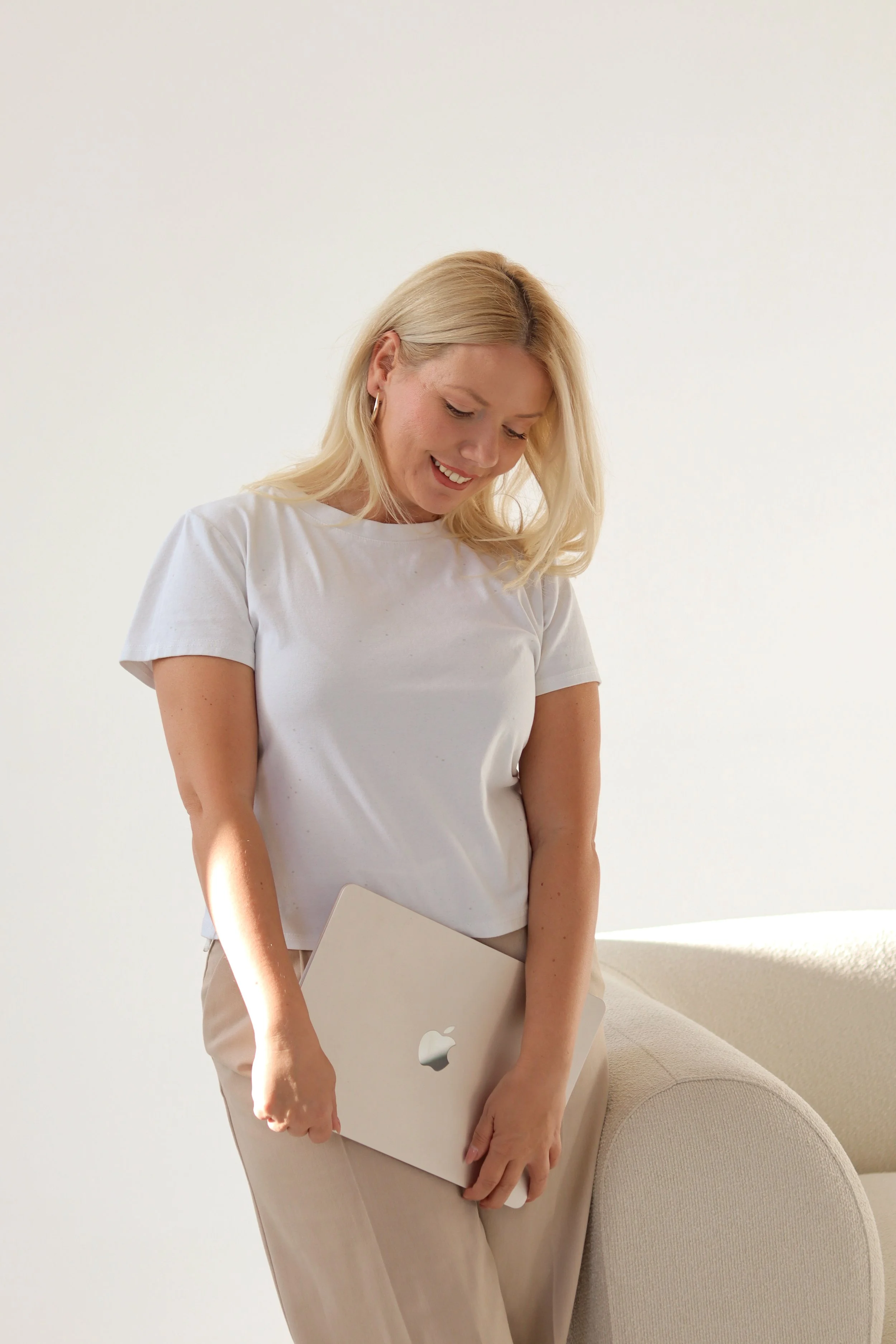 A woman with shoulder-length blonde hair wearing a white t-shirt and beige pants, smiling, holding a closed silver MacBook laptop with an Apple logo, standing in a room with a white wall and beige sofa.