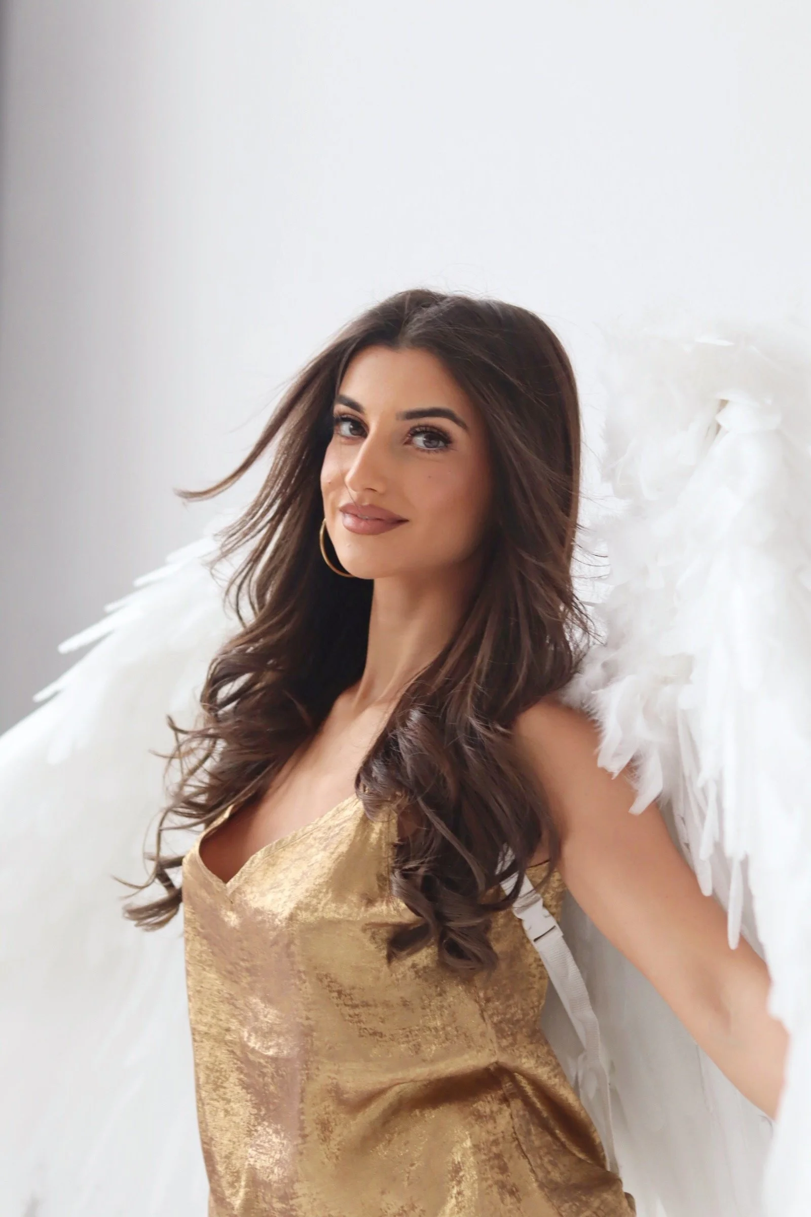 A woman with long brown hair, dressed in a gold dress, wearing large hoop earrings, smiling slightly, with white angel wings behind her, standing against a plain white background.