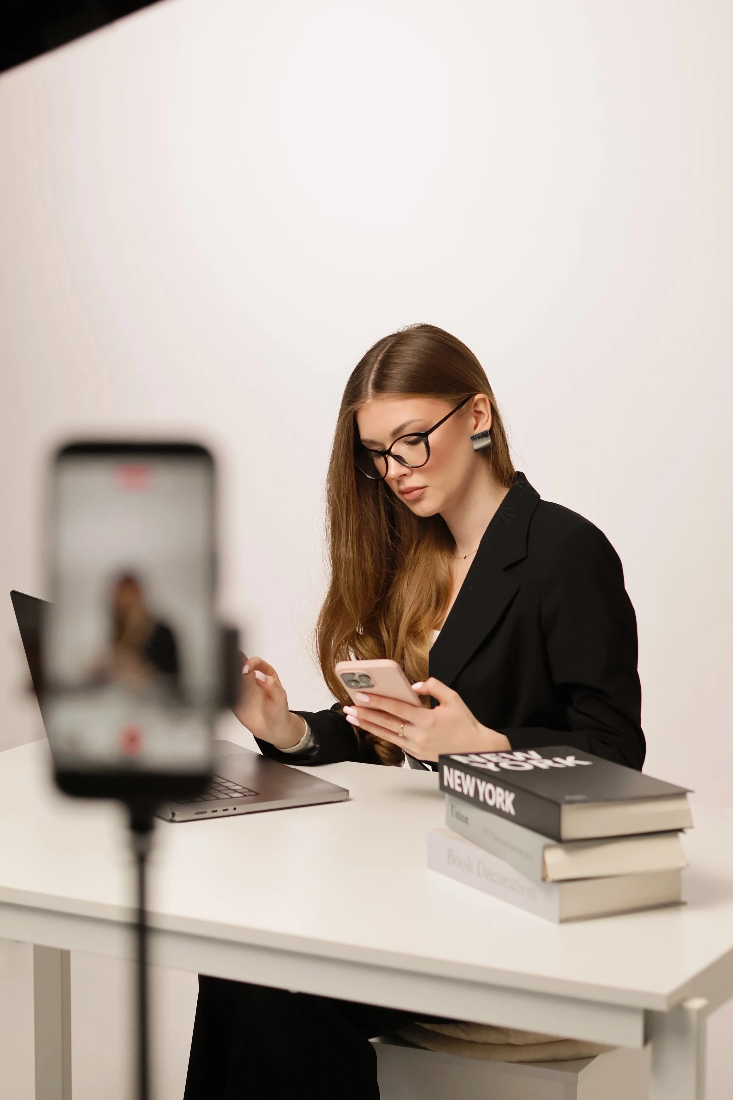 A woman with long brown hair, glasses, and earrings, dressed in a black blazer, is sitting at a desk, using her phone. There are books on the desk titled 'NEW YORK' and 'Book Decorations,' and a laptop. A smartphone on a tripod is in the foreground, partially out of focus.